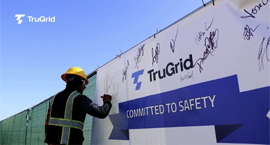 Worker wearing a hard hat and safety vest signing a TruGrid “Committed to Safety” banner covered in team signatures